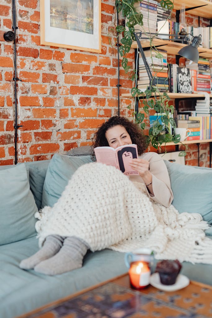 Woman reading on a sofa with a blanket in a cozy room with a brick wall and bookshelves.