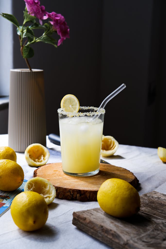 A glass of homemade lemonade with a straw surrounded by fresh lemons and vibrant flowers on a wooden table.